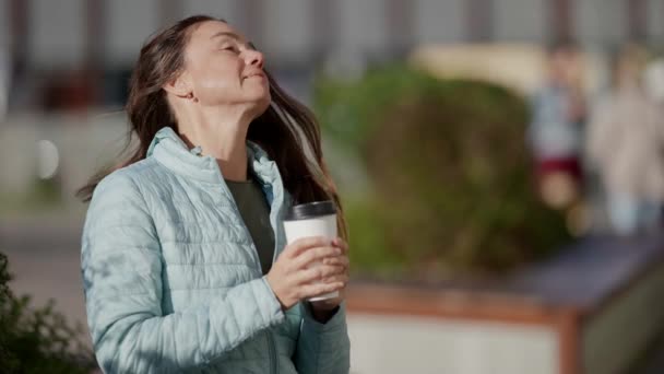 Egy felnőtt, vidám nő, hosszú hajjal, aki egy papírpohárból iszik kávét az utcán. Elvitt italok a kávézóban. — Stock videók