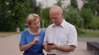 Şehir parkında yürüyüş yapan yaşlı evli bir çift uzak akrabalarıyla iletişim kurmak için telefon ekranına bakar.