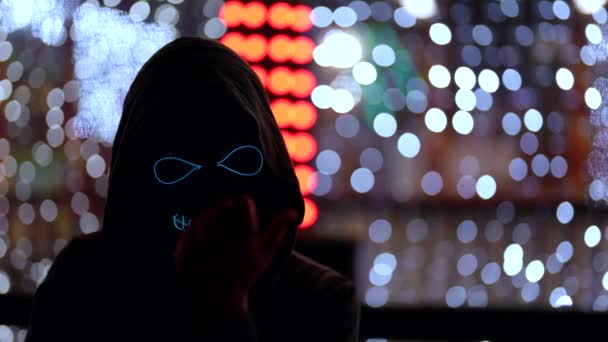 A masked man with glowing eyes and teeth in the image of death beckons with a hand gesture to himself. It stands against the background of neon lights in the dark — Stock Video