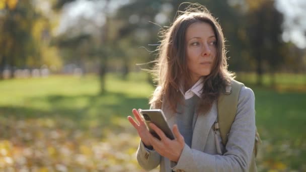 Красивая взрослая туристка с телефоном и рюкзаком стоит в городском парке и пытается найти карту в Интернете для поиска маршрутов — стоковое видео