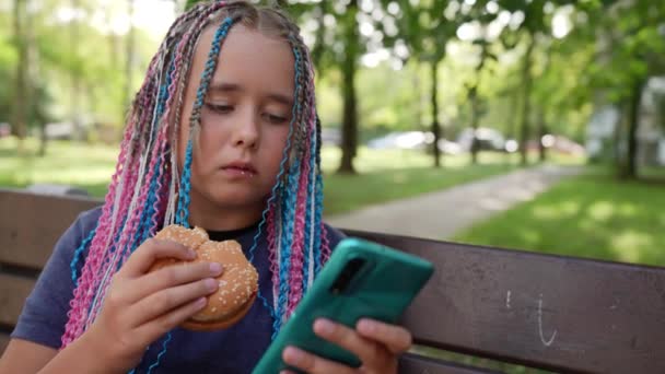 Glad snygg tonåring med afro flätor äter en hamburgare och använder en smartphone på gatan — Stockvideo