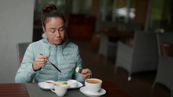 Šťastná dospělá žena sedící v kavárně a pojídající tvarohové koláče s kávou. Ranní jídlo v útulné restauraci ve velkém městě. Víkend v hotelu — Stock video