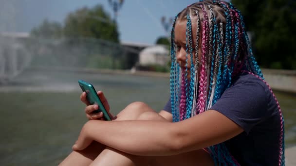Девочка-подросток с афро-косичками держит мобильный смартфон с сенсорным экраном, ходит по улице, солнечный свет на улице — стоковое видео