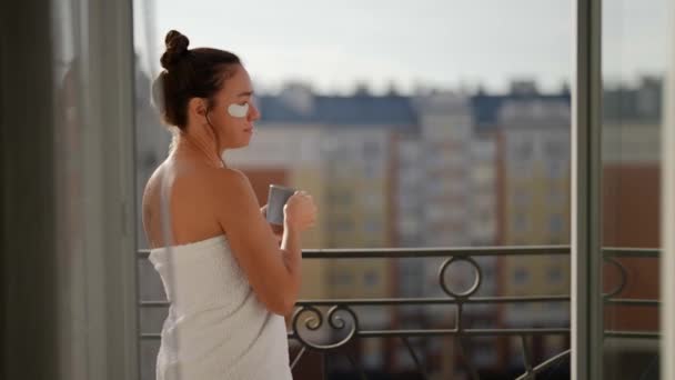 Μια κομψή γυναίκα με λευκή ρόμπα βγαίνει στο μπαλκόνι το πρωί για να πιει καφέ στον καθαρό αέρα. — Αρχείο Βίντεο