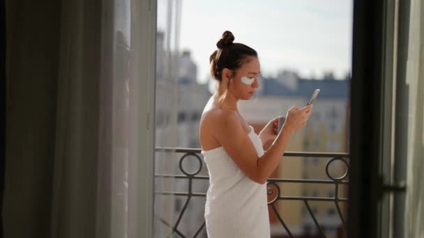 Una mujer adulta con un abrigo blanco con parches en los ojos para proteger la piel envejecida de su cara por la mañana en el balcón con un teléfono para ver las redes sociales y tomar café al aire libre — Vídeo de stock