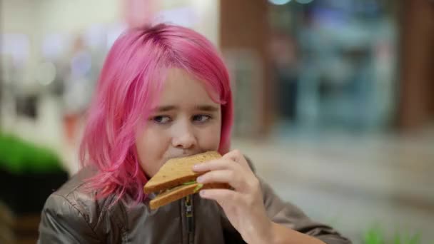 Sorgsen hemlös tonåring med rosa hår äter en smörgås på ett café — Stockvideo