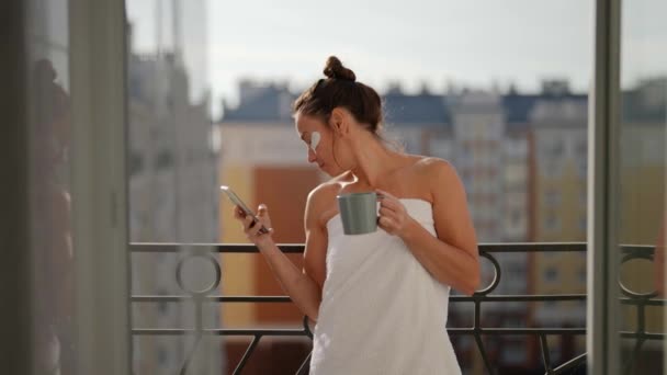 Красивая женщина пьет кофе дома утром, используя смартфон в квартире на чердаке в пижаме — стоковое видео