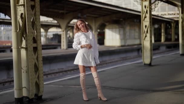 Une jolie jeune femme blonde élancée se tient sur le quai de la gare par une journée ensoleillée en bottes hautes — Video