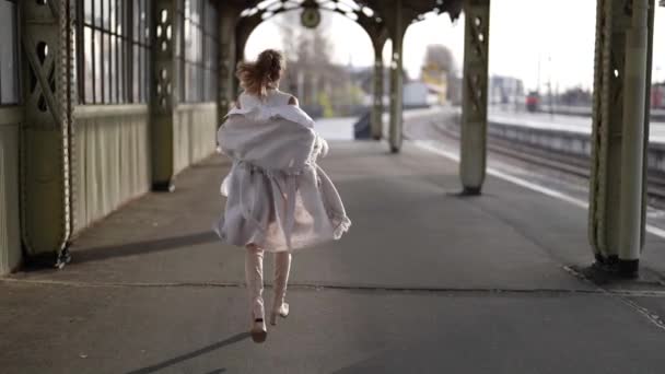 Стройная элегантная блондинка бежит по платформе и размахивает своими шелковистыми волосами на ветру на размытом фоне железнодорожной станции. — стоковое видео