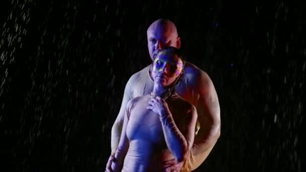 Ett lyckligt vuxet atletiskt par njuter av ösregnet på natten. En kvinna i mask. En kärlekshistoria framförd i neonljus i vattenstrålar. Långsamma rörelser. — Stockvideo