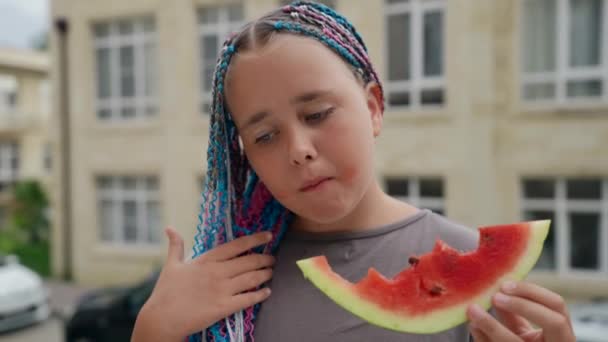 Retrato de cerca de una hermosa adolescente con coletas negras comiendo una fruta de sandía de pie en la calle durante el día — Vídeos de Stock