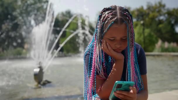Trauriges internetsüchtiges Teenager-Mädchen auf der Straße vor dem Hintergrund eines Brunnens sitzt mit einem Telefon und kommuniziert in sozialen Netzwerken mit Freunden — Stockvideo