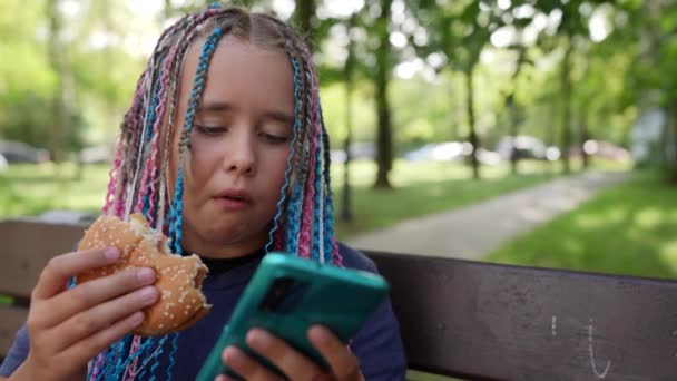 Un adolescente con un teléfono inteligente está comiendo una hamburguesa en la calle. Comida chatarra para colegialas. Comunicación en la red social chica sándwich — Vídeos de Stock