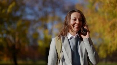 Sıradan kıyafetler giyen ve şehir parkında akıllı telefon kullanan çekici bir yetişkin kadın. Sonbaharda sokakta insanlarla çevrimiçi iletişim kurmak.