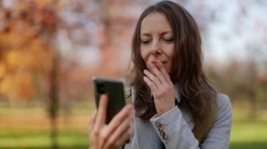 Parkta bir sonbahar günü akıllı telefonun içeriğini kontrol eden çekici bir kadının ön görüntüsü.