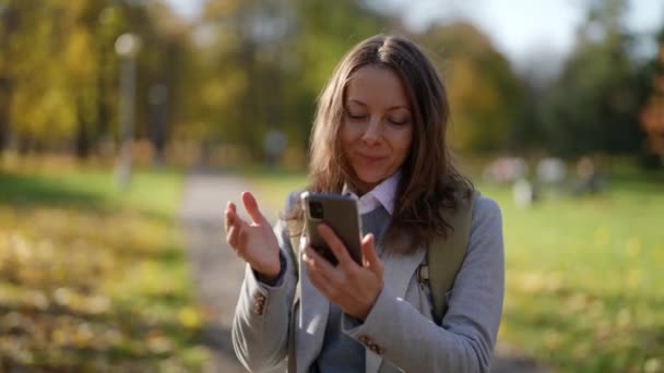 Ritratto di un'attraente donna europea vestita con abiti casual e che utilizza uno smartphone in un parco cittadino. Comunicare con le persone online, scambiare messaggi e navigare in Internet. — Video Stock