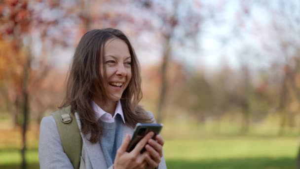 행복 한 성인 여성 이 따뜻 한 가을 화창 한 날 이 공원을 산책하며 핸드폰을 들여다보고 있습니다. 유쾌 한 외모를 하고 있는 성인 여자는 비디오 통화중에 긍정적 인 감정을 받는다. — 비디오