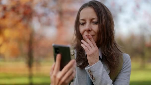 Pandangan depan seorang wanita menawan yang berjalan dengan memeriksa isi smartphone pada hari musim gugur di taman — Stok Video