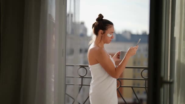 Una hermosa mujer adulta en una habitación de hotel en el balcón bebiendo una taza de café o té viendo las noticias en su teléfono — Vídeo de stock