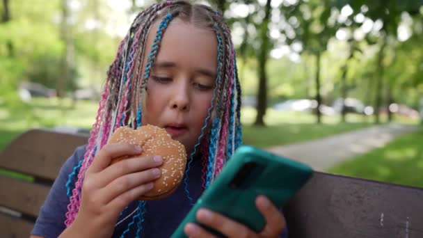 Hungrige Teenie-Mädchen essen einen Burger auf der Straße. Mädchen mit Handy beißt Cheeseburger-Fast-Food — Stockvideo