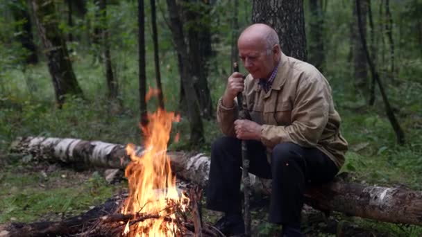 Un vieil homme réfléchi s'assoit seul près d'un feu de camp dans les bois tard dans la nuit, regarde les flammes du feu et se souvient de sa vie, passant son temps libre à l'extérieur, profitant de la solitude. — Video