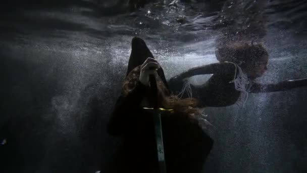 Un hombre misterioso con una espada está sentado en el fondo bajo el agua en un estanque y una mujer en un vestido negro nada por — Vídeo de stock