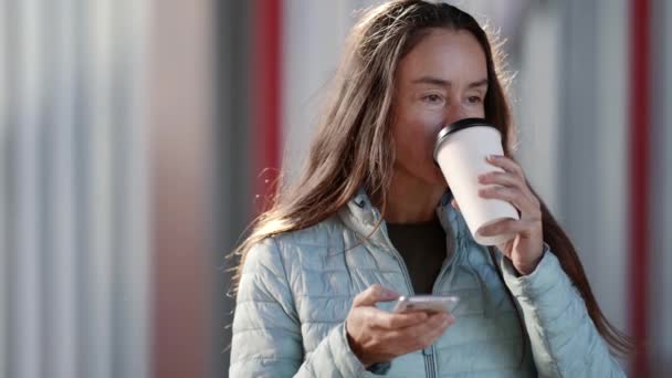 在一个现代化的城市里，一个穿着休闲装的漂亮女人在纸杯里享受着热饮，在互联网上使用着手机 — 图库视频影像