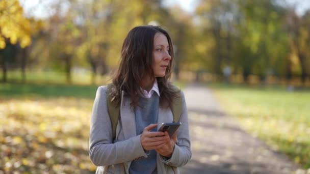 中年女性は秋の週末に公園を歩きスマートフォンでチャットでメッセージを読んでいます — ストック動画