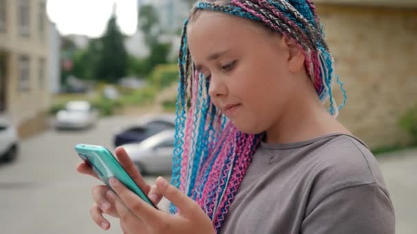 Egy stílusos frizurájú tinédzser lány áll az utcán egy mobiltelefonnal. Kommunikáció, közösségi hálózatok, az online játékok fogalma — Stock videók