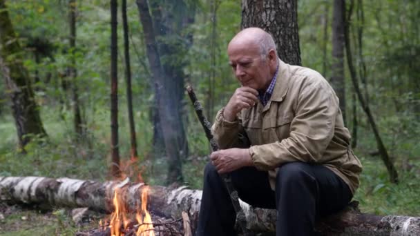 Le vieil homme joyeux repose dans la forêt, regardant le feu ardent, le repos dans la nature — Video