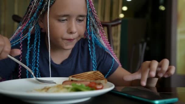 Голодная девочка-подросток ест в кафе и пользуется смартфоном — стоковое видео