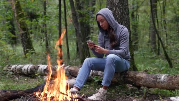 Distanciamento social uma mulher solitária está sentada perto de uma fogueira na floresta e usa seu telefone para se comunicar com a Internet. Passe o seu tempo livre na natureza. Uma mulher desfrutando de sua solidão. — Vídeo de Stock