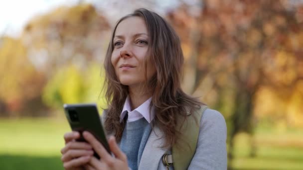 Parkta akıllı telefonlu zeki bir kadın güneşli bir günde yürüyor, okuyor ve mesaj gönderiyor. — Stok video