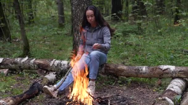 Distanciamento social Uma mulher adulta solitária está sentada perto de uma fogueira em uma floresta de pinheiros. Passe o seu tempo livre na natureza. Uma mulher a desfrutar da solidão. O conceito de recreação — Vídeo de Stock