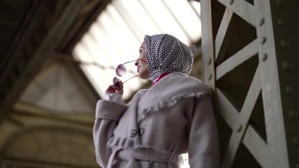 Hermosa mujer en abrigo, pañuelo para la cabeza y gafas de sol está esperando a alguien en la estación — Vídeos de Stock