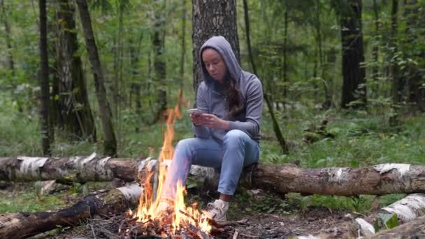 Mujer con teléfono inteligente en el bosque, sentado cerca de fogata y charlando en línea por las redes sociales — Vídeo de stock