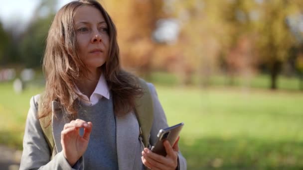 Akıllı telefonlu bir kadın parkta el sallayıp arkadaşlarıyla selamlaşan birini bekliyor. — Stok video