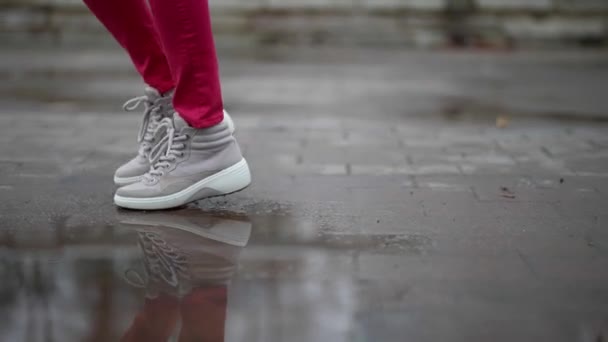 Yolda yürürken, su birikintilerinde mutlu bir şekilde zıplayan bir kadının bacakları. Spor ayakkabılı bir kadın sokakta yürüyor. Şehirde yürüyen kendine güvenen bir kızın bacakları.. — Stok video
