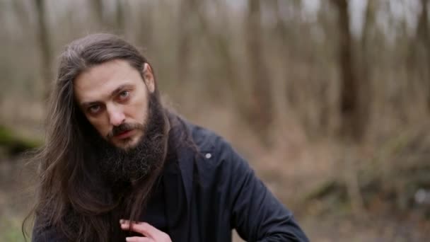 Загадочный человек с бородой и длинными волосами одет в черную одежду, загадочно глядя в камеру — стоковое видео