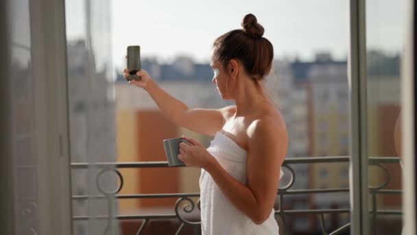 A gorgeous adult woman stays on the balcony, holds a cup of hot coffee or tea, admiring the beautiful city view, takes pictures of herself on the phone and enjoys relaxing — стоковое видео
