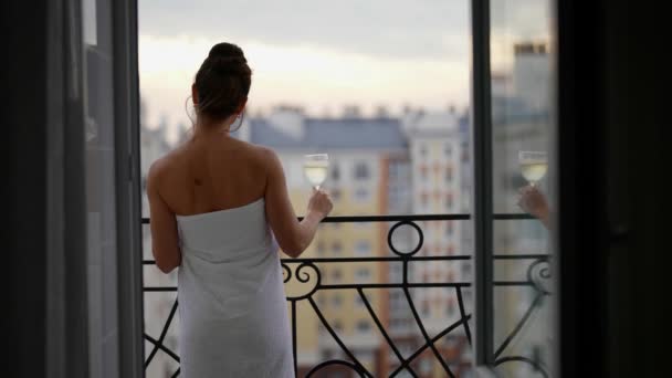 Een slanke volwassen vrouw in de avond na een douche in een handdoek staat op het balkon met een glas champagne en geniet van het stadsgezicht — Stockvideo