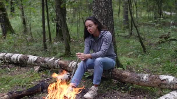 An adult woman enjoying her solitude in the forest during a hike is warming herself by the fire — Stockvideo