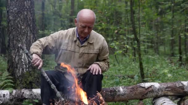 A man at a campfire in the forest. An old man looks at a forest fire in nature in summer. A lonely traveler is sitting and resting by the campfire — Stock videók
