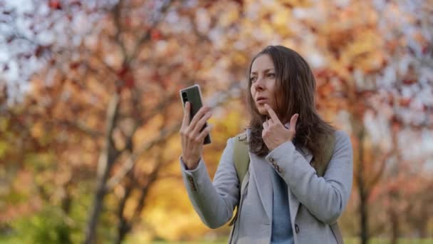 Adult woman is calling by video conference in smartphone, walking in park in sunny fall day — стоковое видео