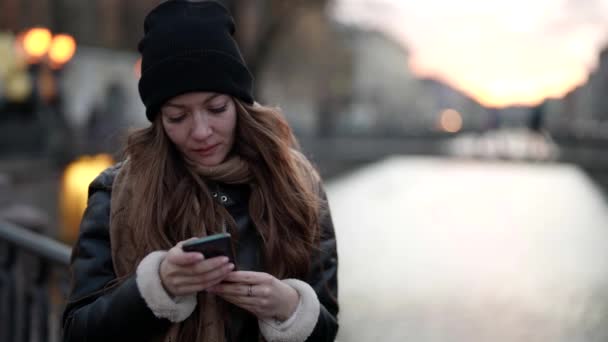 Mujer joven reflexiva con teléfono inteligente en el puente del pequeño río en la ciudad, mensaje de lectura — Vídeo de stock