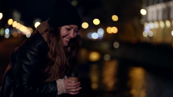 Mujer alegre con taza de café para ir en terraplén de la ciudad en la noche — Vídeos de Stock