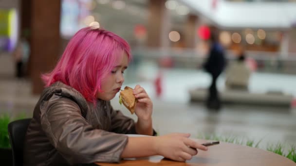 Pembe saçlı genç bir kız alışveriş merkezindeki bir kafede ekmek ve sosis yiyor ve mobil bir uygulamaya bakıyor. İşlevsiz bir aileden bir çocuk. — Stok video