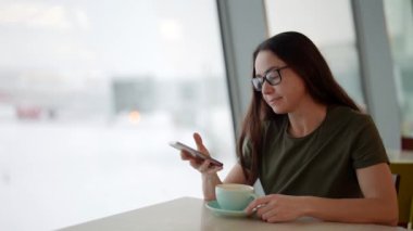 Yetişkin bir kadın havaalanı kafesinde dinleniyor, kahve içiyor ve akıllı telefon kullanıyor.