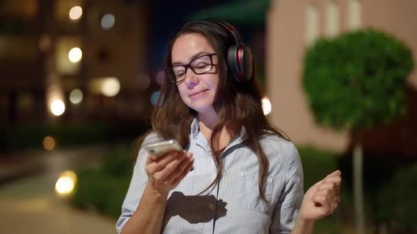 Mujer feliz con auriculares está escuchando música en la calle de la ciudad en la noche, utilizando el teléfono inteligente — Vídeos de Stock