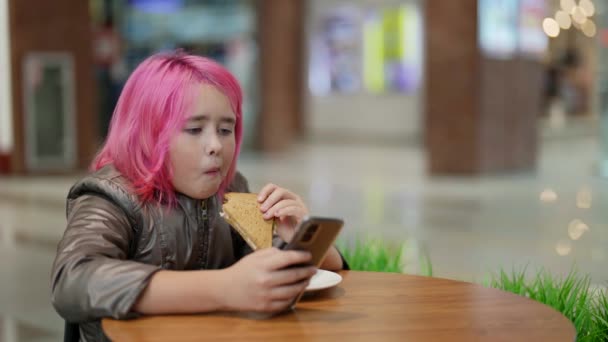 Waif nastolatka z różowymi włosami jedząc chleb i kiełbasę w centrum handlowym kawiarni i przeglądając aplikację mobilną — Wideo stockowe